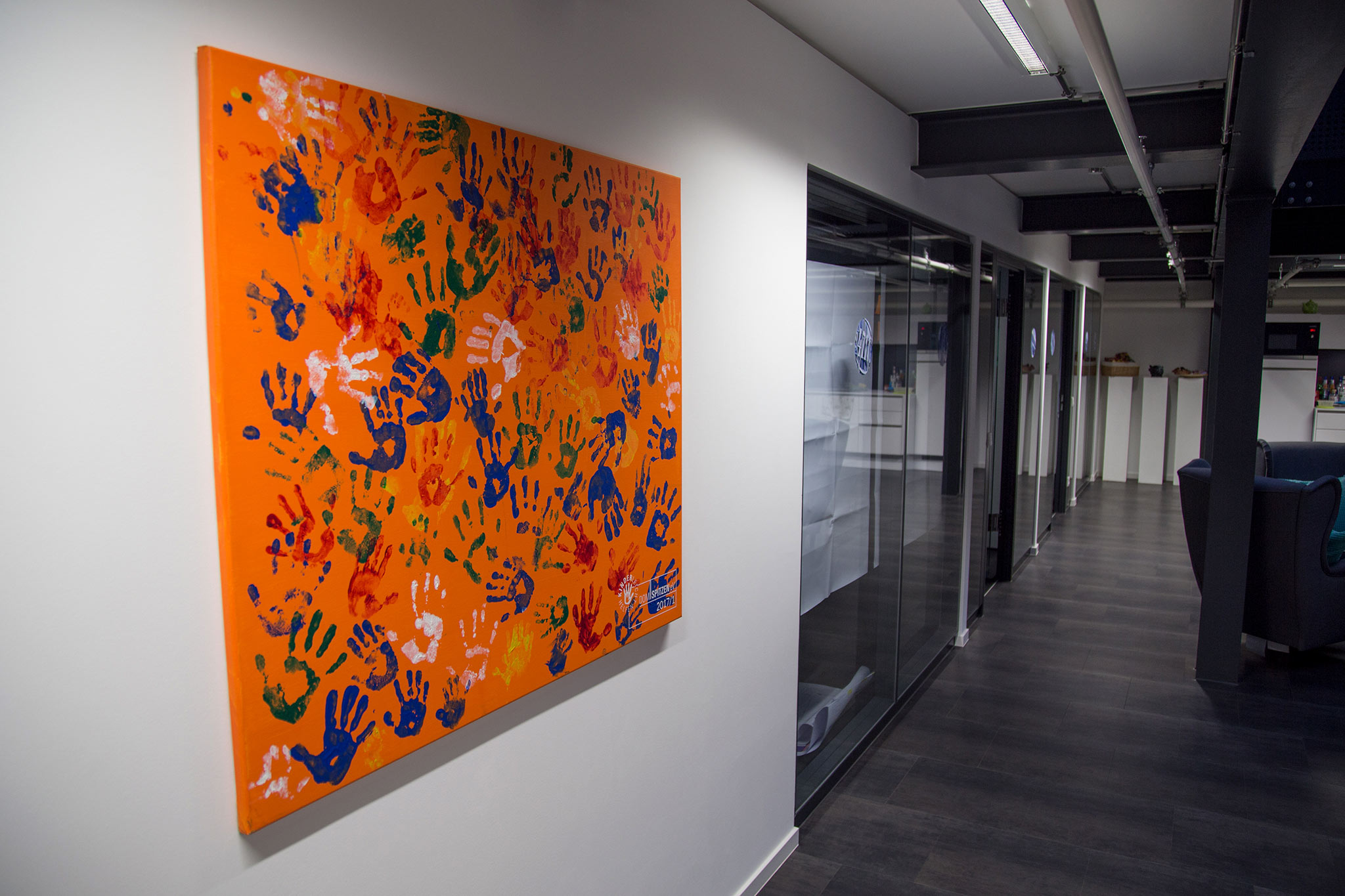 Ein Bild (orangener Hintergrund mit bunten abdrücken von Kinderhänden) hängt im Büro der Agentur Wunderman. Domspitzen eV.